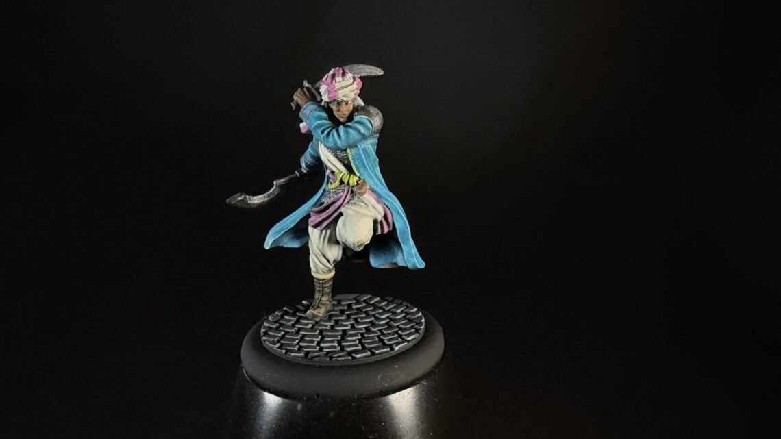 Tabletop miniature of a pirate-like fighter in a blue coat, pink scarf, and white pants, posed with a curved blade, standing on a cobblestone base against a dark background